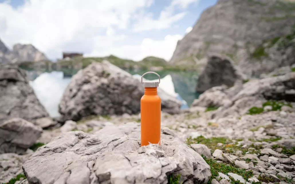 A sturdy orange water bottle sitting on a rock in the mountains.